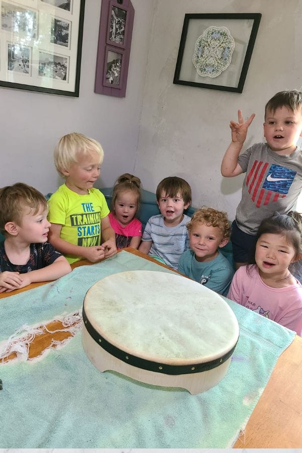 A group of children gather around a table with a large drum, in a room adorned with framed art, eagerly exploring lesson plans on Irish culture.