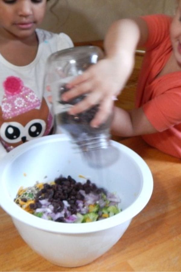 Two children add ingredients to a bowl on a table, exploring exciting broccoli recipes for kids. One child pours from a jar, mixing in chopped vegetables and black beans, creating a colorful creation.