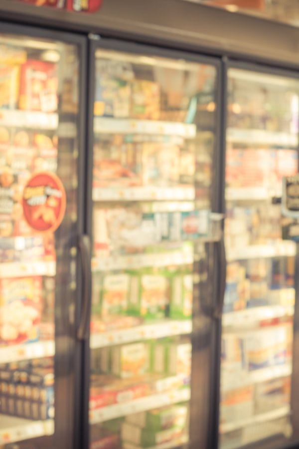 A blurred image of a supermarket aisle showcases refrigerated glass doors filled with various packaged food items, some prominently displaying CN Labels for CACFP compliance.