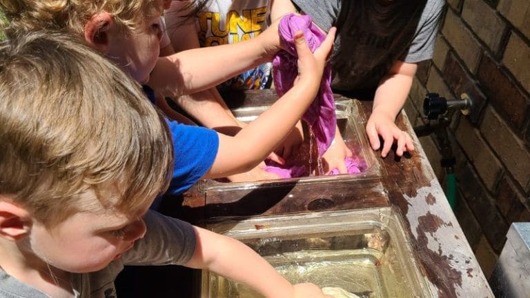 Children joyfully playing with water and natural dyes in fabric-filled trays outdoors.