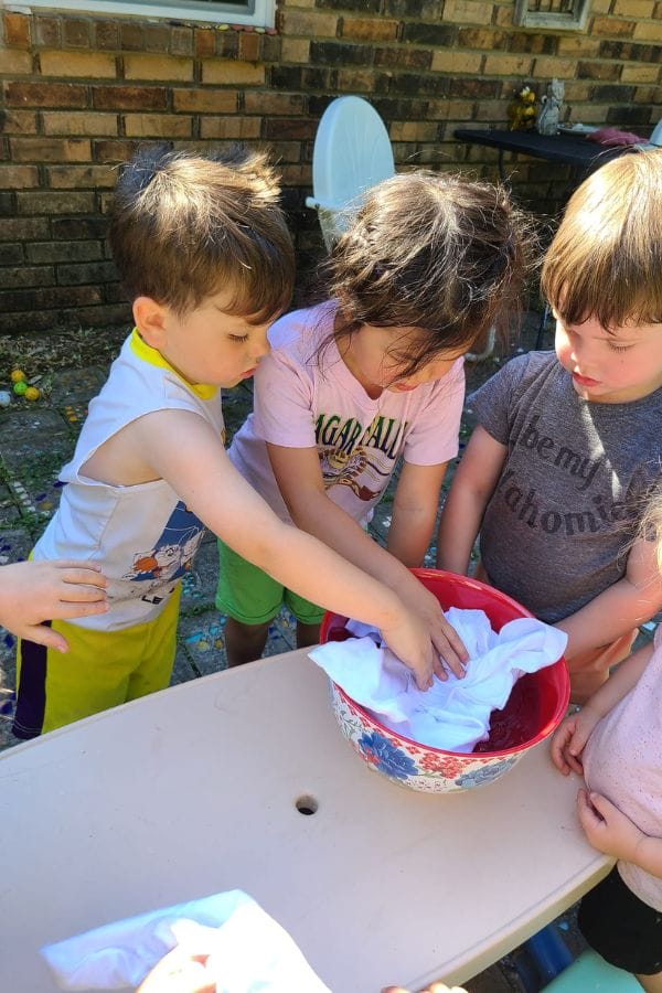 Three children gather around a table, using natural dyes to color fabric in a red and white bowl outdoors.