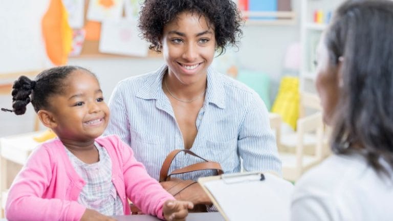 A woman and a smiling child sit across from another person holding a clipboard in a classroom setting, where the open door policy fosters an atmosphere of transparency and communication.
