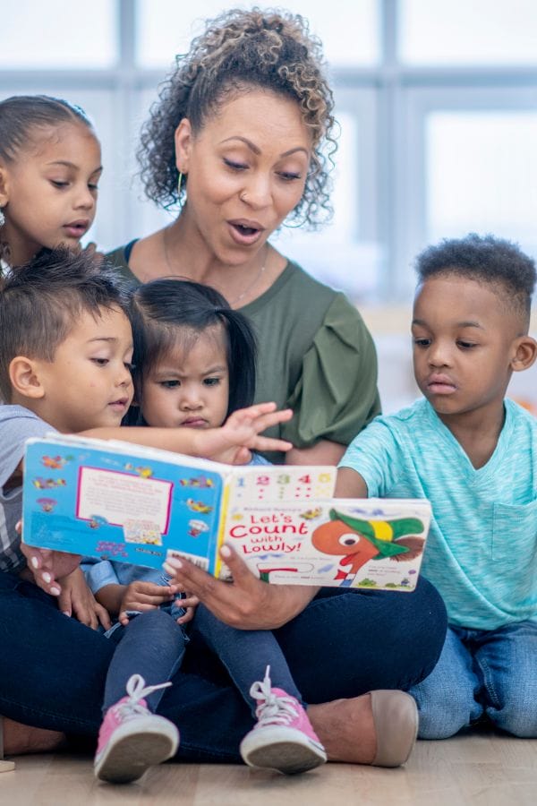 A woman sits reading a colorful book to four young children, gathered around her like an open door, welcoming their curious eyes and attentive minds to the vibrant stories on the pages.