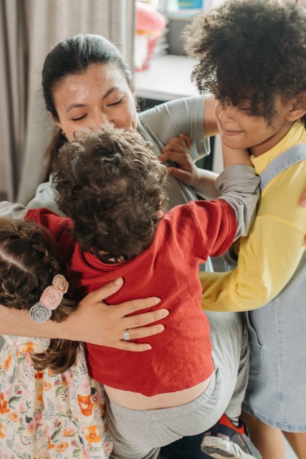 A woman embraces three children in a joyful group hug indoors, embodying the warmth of an open door policy that invites love and connection.