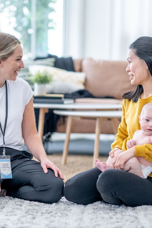 A woman in a white shirt engages warmly with a woman in a yellow sweater, who is holding a baby, as they sit comfortably on the living room floor. Their easy conversation reflects an open door policy that fills the home with welcome and joy.
