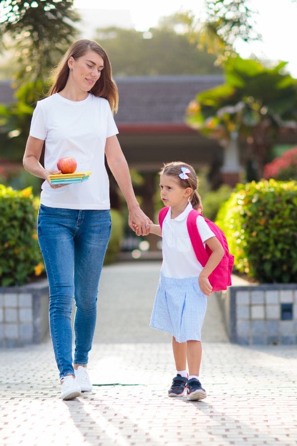 A woman holding books and an apple walks hand-in-hand with a young girl wearing a pink backpack, embodying an open-door policy to learning. They are outdoors, surrounded by the lush embrace of nature.