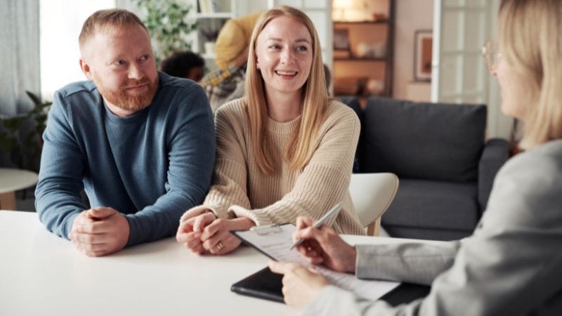 A couple sits at a table across from a professional, engaged in effective communication, with a clipboard visible in the foreground.