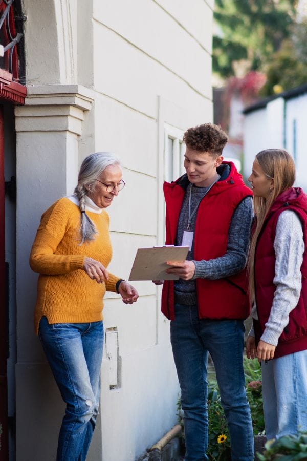 Three people are engaged in effective communication outside a building: an older woman in a yellow sweater and two younger individuals in red vests, one holding a clipboard.
