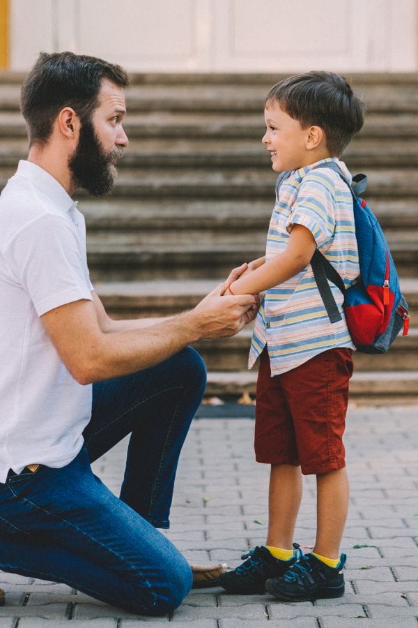A man kneels on one knee, engaging in effective communication as he holds hands with a smiling child wearing a backpack, framed by the steps behind them.
