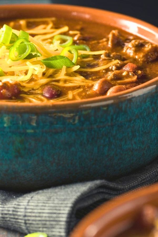 A bowl of chili, perfect for picky eaters, is topped with shredded cheese and sliced green onions, nestled in a blue ceramic dish. A delightful addition to your crockpot recipes collection.