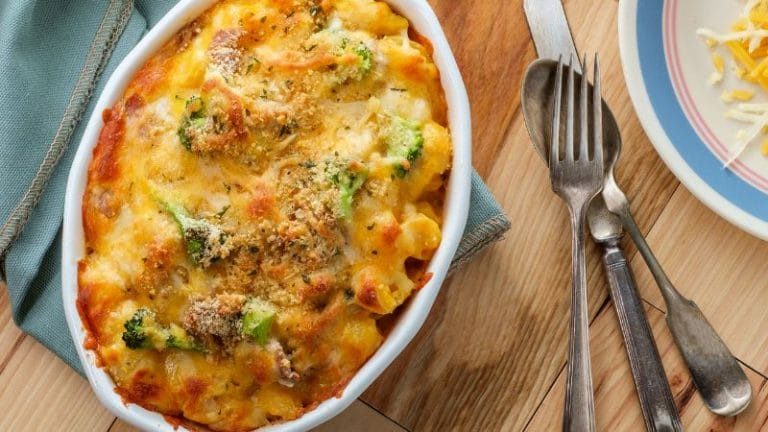 A simple tuna casserole with cheese, broccoli, and breadcrumbs sits in an oval dish on a wooden table. Beside it, a blue napkin and three forks await the meal.