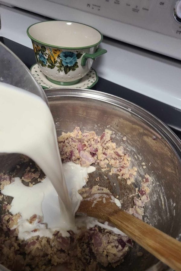 Milk is being poured into a bowl with a mixture of ingredients, including diced onions, reminiscent of preparing a Tuna Rice Casserole. A floral cup sits on a stove in the background, adding charm to the kitchen scene.