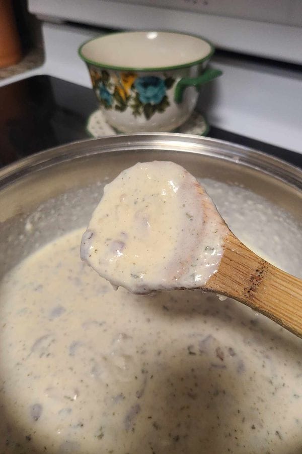 A wooden spoon holds a creamy white sauce, rich with herbs and small chunks, hinting at the heartiness of a tuna rice casserole being prepared in the pot. A floral mug sits in the background, completing this cozy kitchen scene.