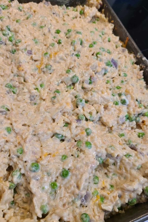 A close-up of a creamy tuna rice casserole mixture with visible peas, rice, and chopped vegetables in a baking tray.