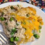 A slice of tuna rice casserole with peas and melted cheese rests on a white plate, next to a fork. The background features colorful, comic-style fabric.