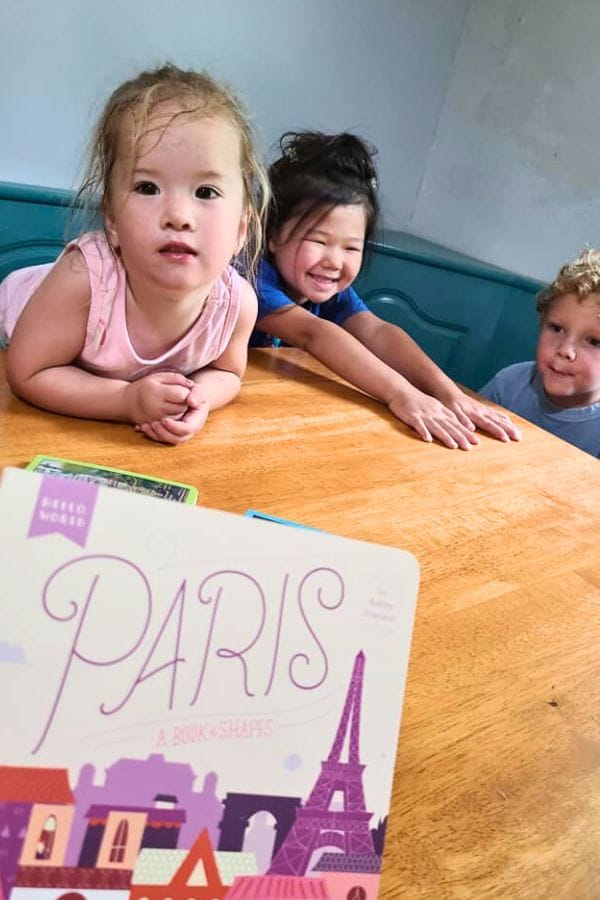 Three young children sit at a table with "Paris: A Book of Shapes" in the foreground, diving into lesson plans on French culture.
