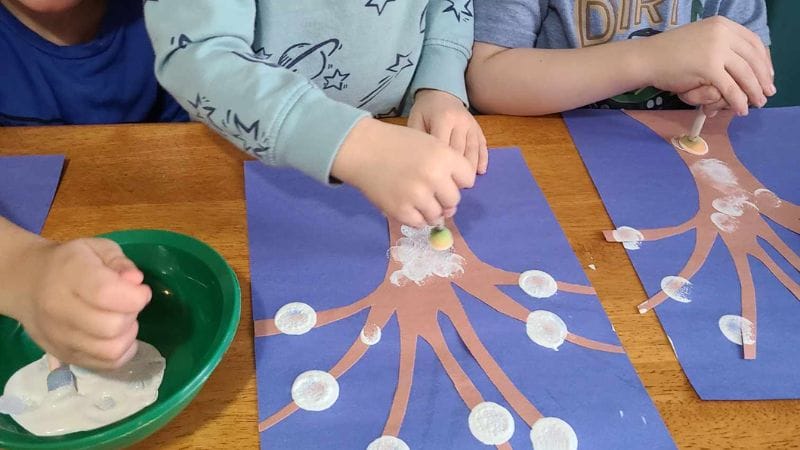 Children creatively paint cherry blossom trees on blue paper, a delightful activity often included in lesson plans on winter to explore seasonal changes through art.