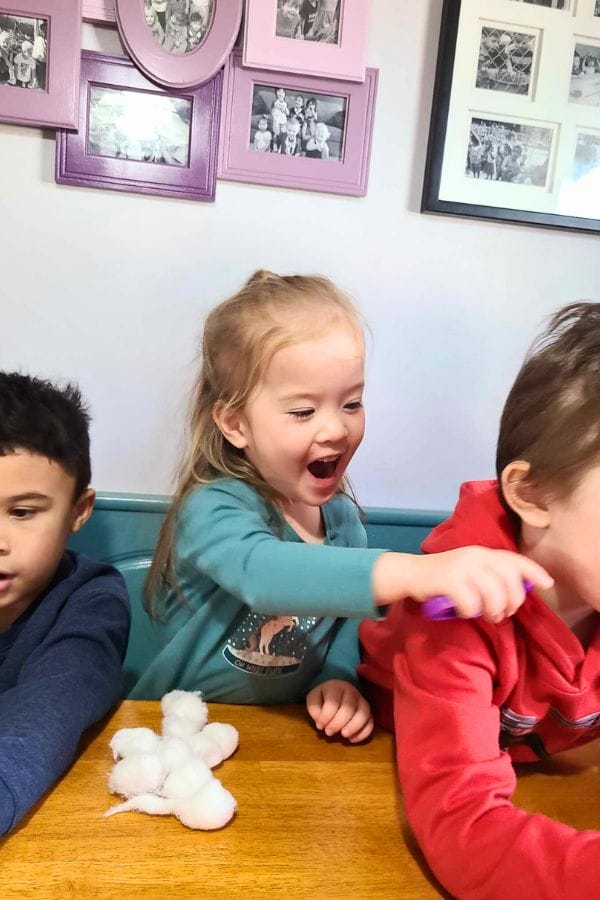 Three children sit at a wooden table, surrounded by the warmth of lesson plans on winter. The girl in the middle reaches out with a smile, while the boy to her right dons a red shirt. A stuffed toy lies on the table, adding charm to their cozy gathering.