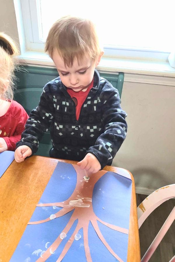 A child sits at a table, engaged in a winter-themed lesson plan, pressing white paint onto a paper tree with their finger.