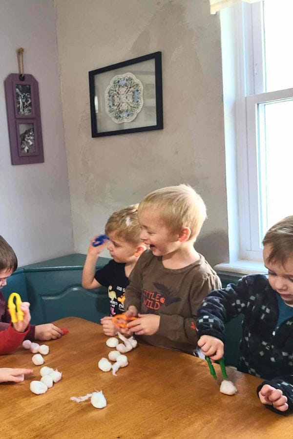 Four children sit at a table near a window, eagerly playing with cotton balls and plastic tweezers. Through this playful activity, they explore elements of Lesson Plans on Winter, sparking creativity and learning as they engage in hands-on fun.