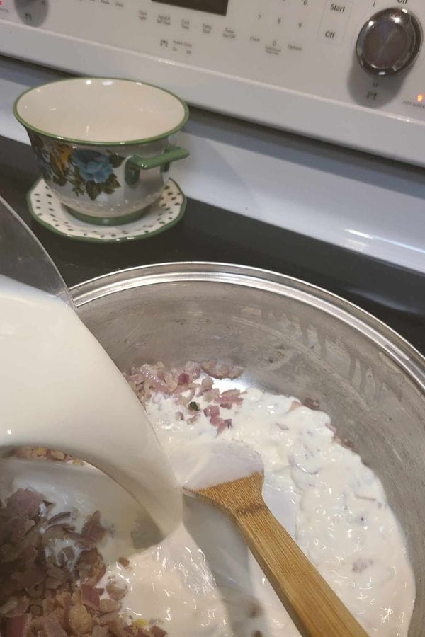 Milk is being poured into a pot with chopped onions, sour cream, and bacon, all stirred with a wooden spatula. A floral cup and saucer rest on the stove beside a can of savory tuna.