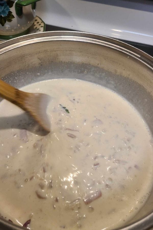 A wooden spoon stirs a rich, creamy white sauce with onions in a pot, infusing it with the savory essence of sour cream and onion tuna.