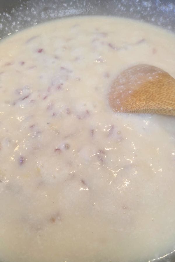 A wooden spoon stirs a creamy, white mixture with small red bits in a pot, resembling the comforting beginnings of a tuna biscuit casserole.