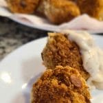 A close-up of two easy tuna croquettes on a plate, one topped with white sauce. More croquettes are visible on a paper towel in the background, showcasing their golden, crispy perfection.