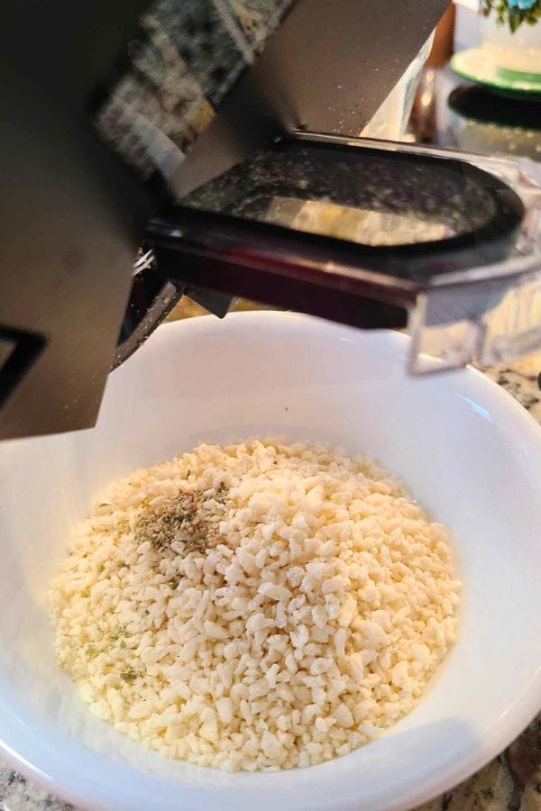 A food processor effortlessly grating cheese into a white bowl on the kitchen counter, perfect for adding a delicious touch to your easy tuna croquettes.