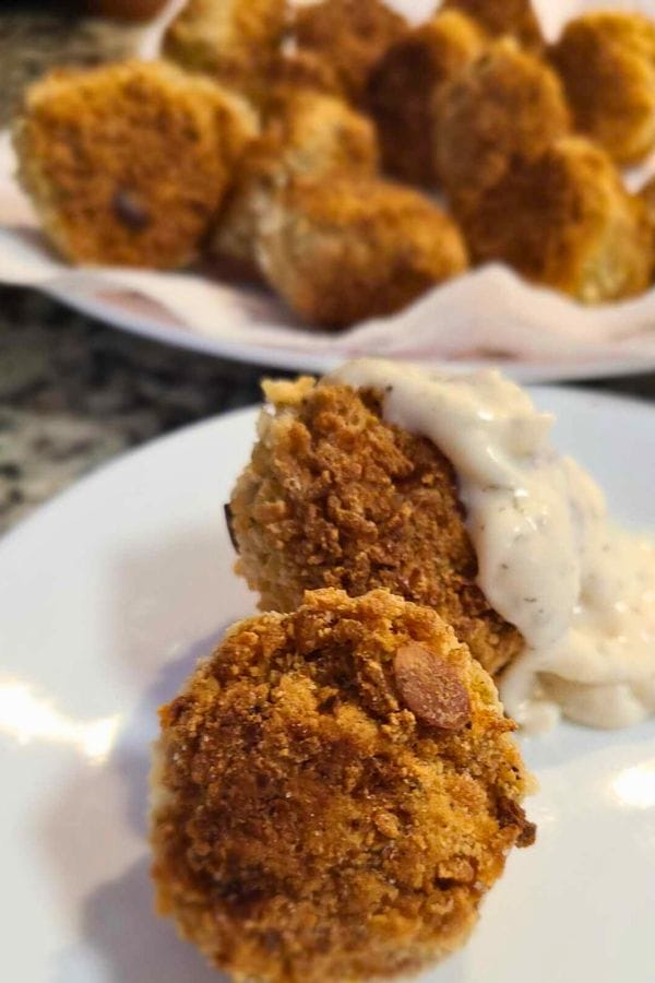 A close-up of a breaded mushroom on a white plate, topped with creamy sauce, evokes the delightful simplicity of easy tuna croquettes. In the background, a blurred plate holds more tempting mushrooms.