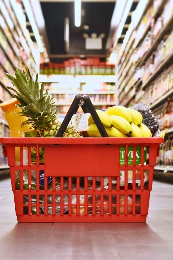 A red shopping basket brimming with bananas, pineapple, and various groceries sits on the supermarket aisle floor. Perfect for those crafting a balanced daycare meal plan template.