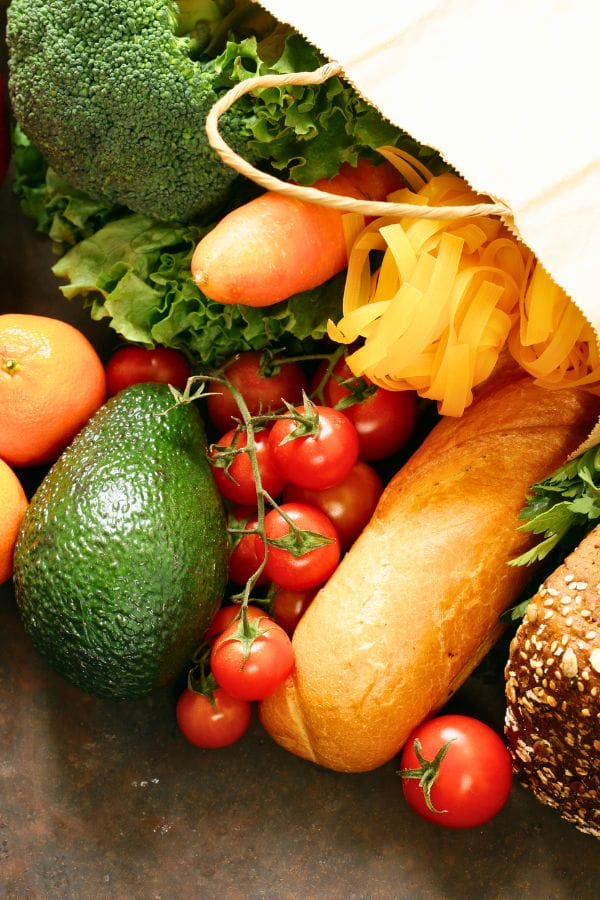 A grocery bag brimming with broccoli, lettuce, carrots, tomatoes, avocado, and more sits on a dark surface—ideal ingredients for crafting a colorful daycare meal plan template.