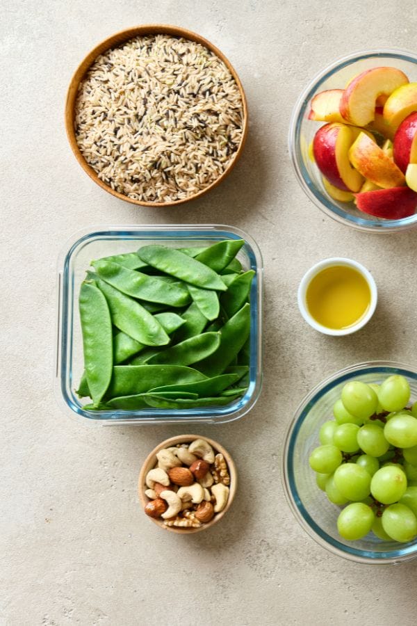 Colorful daycare meal plan template: bowls filled with wild rice, apple slices, snow peas, mixed nuts, and green grapes grace a light surface. A small bowl of olive oil completes this nutritious spread designed to delight little palates.