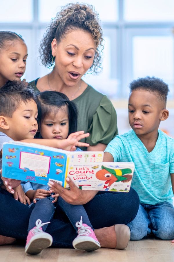 A woman joyfully reads a colorful book to four young children seated around her, showcasing the engaging atmosphere essential for learning. This scene highlights how to advertise a daycare that fosters curiosity and enthusiasm in early education.