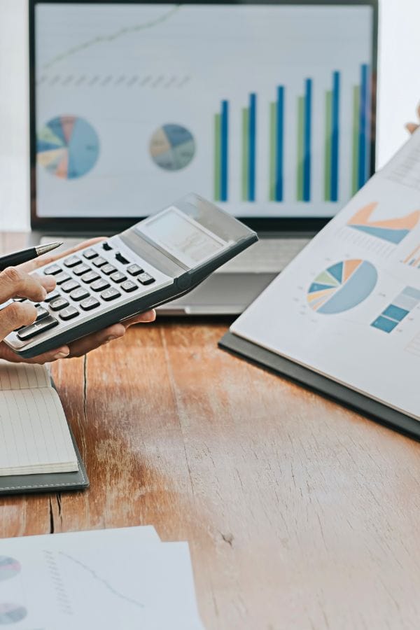 A person is analyzing a financial report with charts while using a calculator. In the background, on a wooden table, a laptop displays graphs. This setting resembles planning strategies akin to learning how to advertise a daycare effectively.