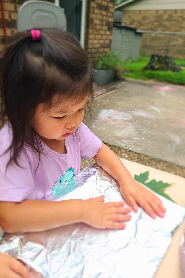 A young girl in a purple shirt engages in leaf rubbing, pressing a vibrant leaf onto foil at an outdoor table. Chalk drawings add a colorful backdrop on the pavement.