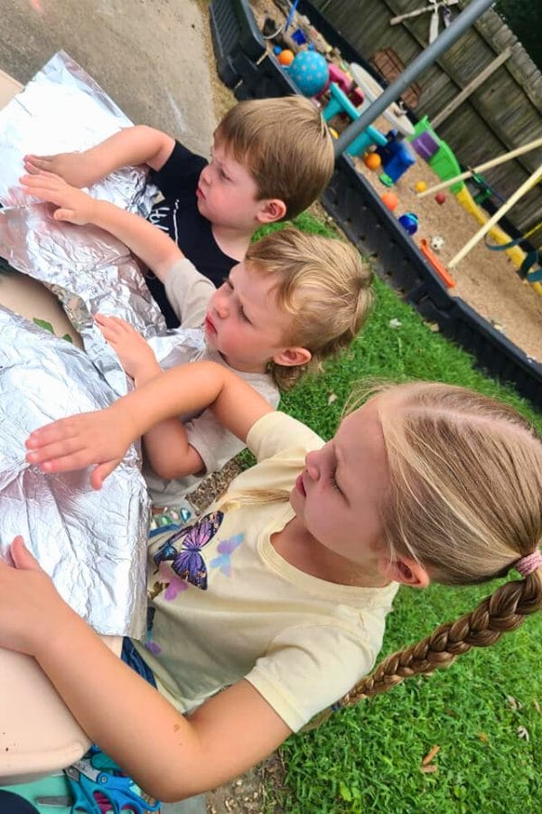 Three children sit at a table outdoors, holding and examining sheets of aluminum foil for their leaf rubbing project, with playground equipment visible in the background.