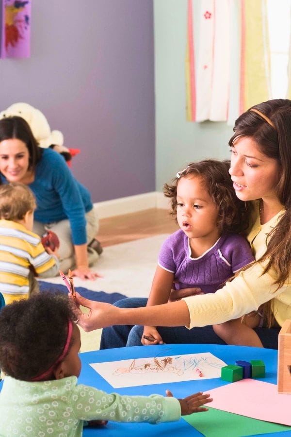 In a vibrant room buzzing with creativity, a caregiver introduces crayons to two children at a table filled with paper and blocks. Nearby, another adult engages a child, embodying the nurturing atmosphere often explored in interview questions for daycare environments.