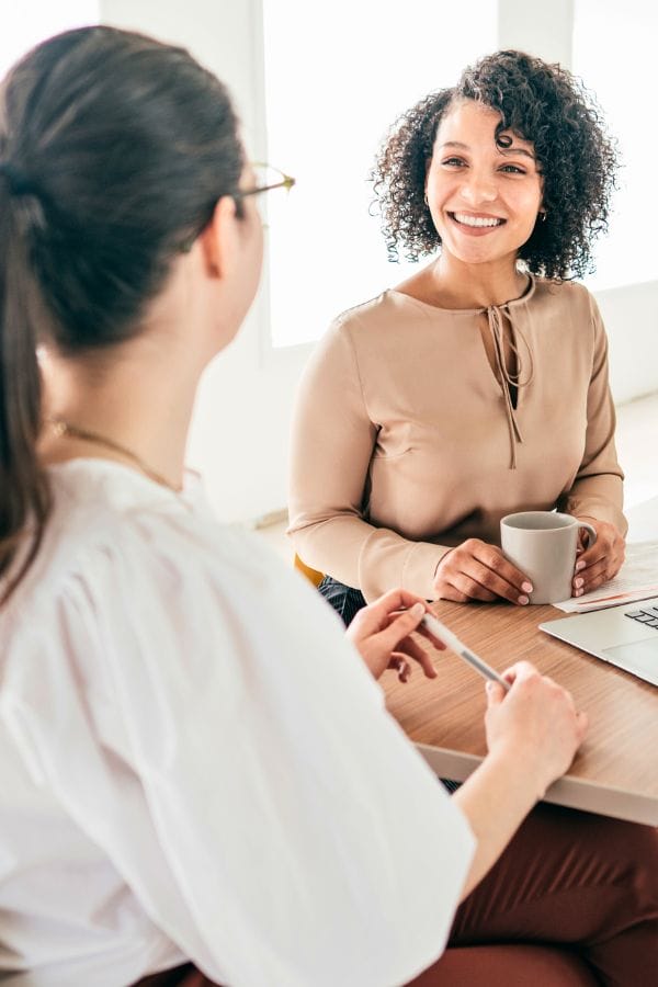 Two women sitting at a table engage in conversation. One holds a mug, while the other, with a pen in hand, jots down notes. A laptop is open in the foreground, perhaps displaying interview questions for daycare roles.
