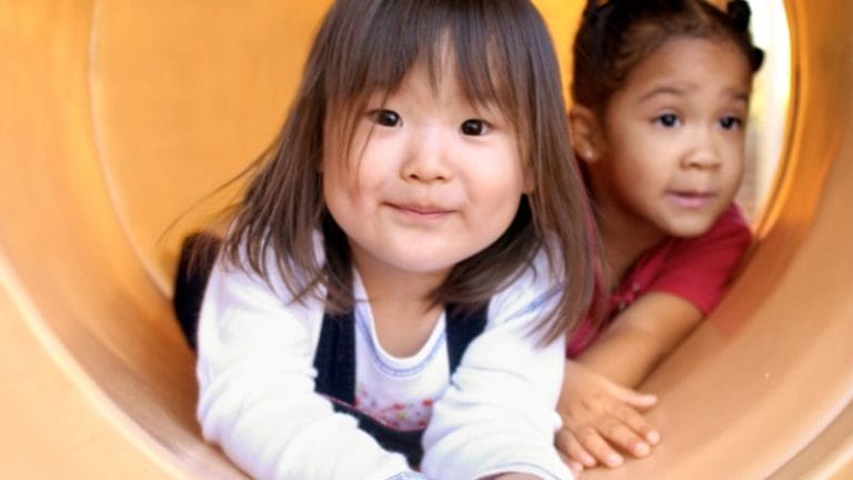 Two young girls are in an outdoor playground tunnel. The girl in the foreground is smiling and looking forward, while the girl behind her looks to the side.