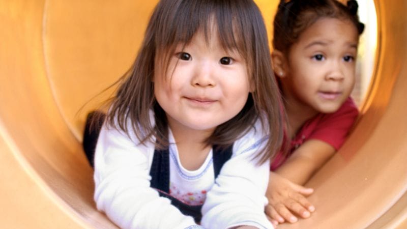 Two young girls are in an outdoor playground tunnel. The girl in the foreground is smiling and looking forward, while the girl behind her looks to the side.