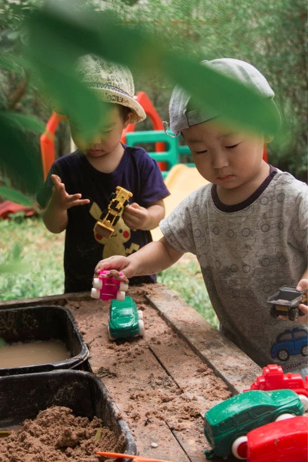 Two children wearing hats play with toy vehicles and sand at a wooden table in the outdoor playground. Green foliage is visible in the foreground.