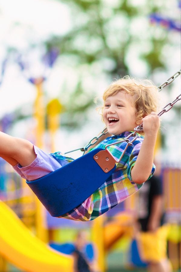 A child with curly hair beams with joy while swinging on a blue swing, enjoying the thrill of outdoor playground equipment.