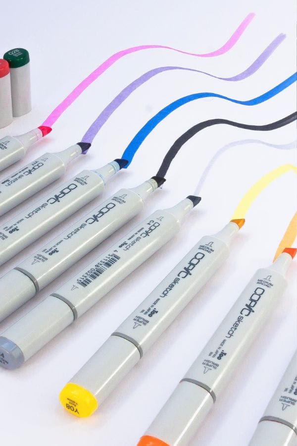 Markers with colored caps (green, red, pink, purple, blue, black, orange, yellow) are arranged on paper, each drawing a wavy line of matching color. These are the best markers for Shrinky Dinks enthusiasts seeking vibrant and consistent hues.