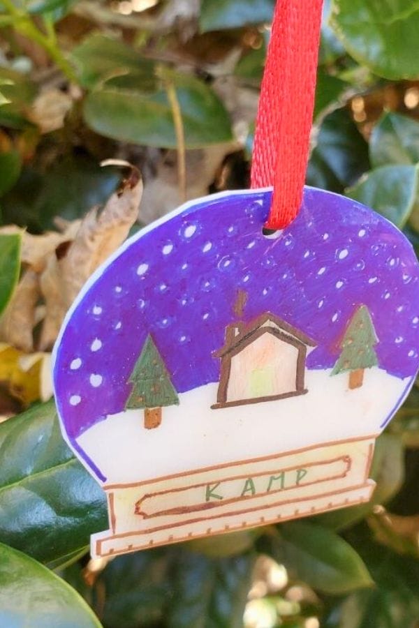 A homemade ornament, crafted with the best markers for Shrinky Dinks, showcases a snowy scene with a house and trees under a starry sky. It hangs on green foliage, adorned with a red ribbon and labeled "KAMP.