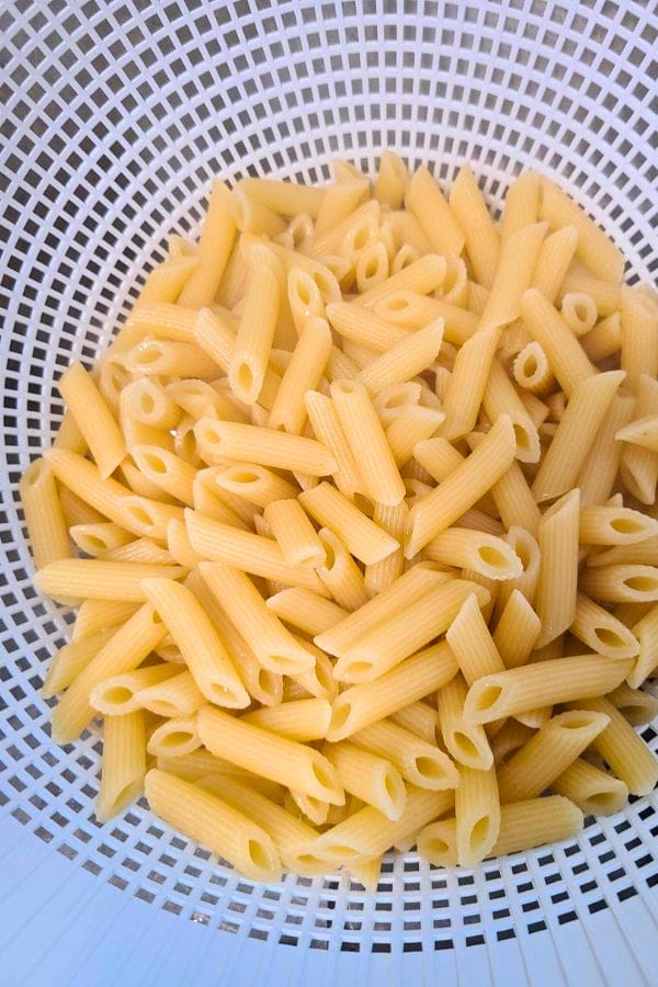 Cooked penne pasta draining in a white plastic colander, soon to be transformed into a delicious Ranch Tuna Pasta Salad.