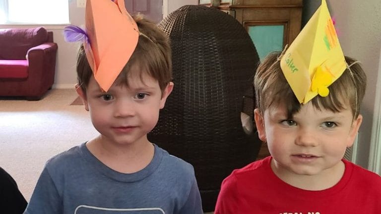Two young boys indoors proudly wear paper hats, standing side by side. With one in a blue shirt and the other in a red shirt, they capture the essence of playful traditions reminiscent of vibrant German culture.