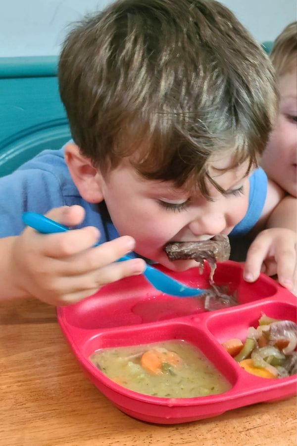A child in a blue shirt enjoys hearty soup and bread from a red divided plate with a blue spoon, reminiscent of a cozy meal steeped in German culture.
