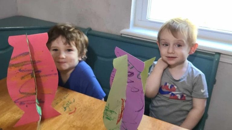 Two children sit at a table adorned with colorful, fish-shaped paper crafts reminiscent of Henri Matisse's vibrant style. A window quietly frames the scene in the background.