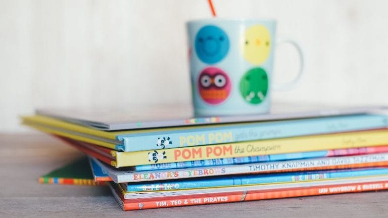 A stack of colorful children's books, including "The Very Busy Spider," rests on a wooden surface, topped with a mug featuring emoji designs and a straw.